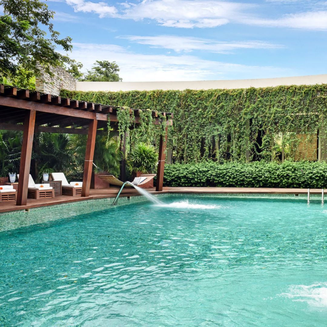 Image Rosewood Mayakoba's Spa Serenity Pool: clear blue water sparkeling in the sun. Pool is surrounded by white lounge chairs, white umbrellas, lush greenery, and planters. There are two fountains at the edge of the pool.
